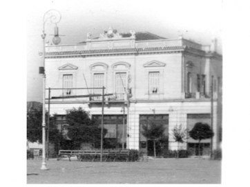 Το μέγαρο Νικ. Καρανίκα στη δυτική πλευρά της πλατείας Θέμιδος.  Στις τρεις πόρτες αριστερά στο ισόγειο στεγαζόταν το ζαχαροπλαστείο  «Βιεννέζικον». Λεπτομέρεια από το επιστολικό δελτάριο αρ. 2  του Φραγκούλη Καλουτά από την Ερμούπολη της Σύρου. Περίπου 1915.  Αρχείο Φωτοθήκης Λάρισας.